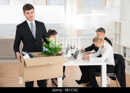 Unglücklich Geschäftsmann die Habseligkeiten in Feld, während andere Leute arbeiten im Büro Stockfoto