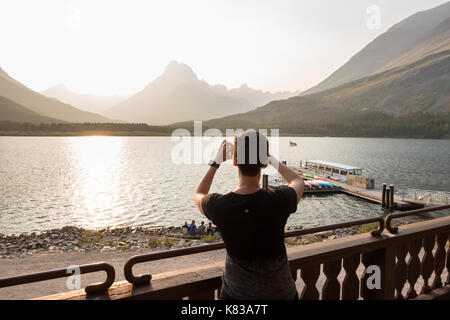 Weibliche Tourist, der ein Foto des Swiftcurrent Lake an vielen Glacier National Park mit dem historischen Chief zwei Pistolen Boot angedockt Stockfoto