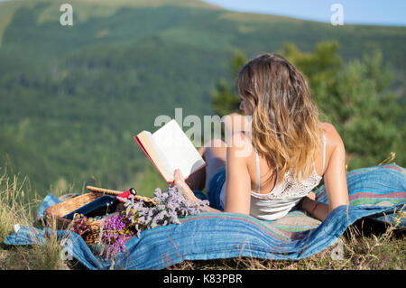 Junge Frau Festlegung auf eine Decke und ein Buch auf einem Picknick im Feld mit Blick auf die Berge Stockfoto