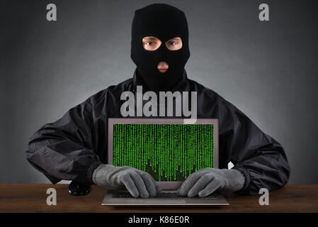 Hacker in schwarze Maske eingeben Auf Laptop mit binären Code Stockfoto
