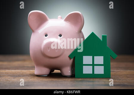 Nahaufnahme des Grünbuchs Haus und Sparschwein auf hölzernen Tisch gegen grauer Hintergrund Stockfoto