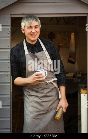 Ein Handwerker das Tragen einer Schürze steht in der Tür eines Workshops, Becher und kleine Log aus Holz, lächelnd. Kaffee Pause. Stockfoto