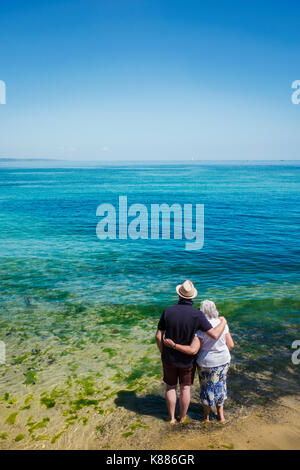 Ansicht der Rückseite des älteren Paares stehend mit Arme um die Schultern aus Blick auf das Meer über die lebendige klaren blauen Wasser. Stockfoto