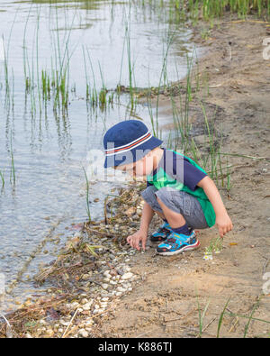 Kleiner Junge sammelt Steine am Rand von einem See oder Teich. Stockfoto