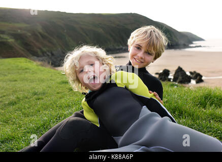 Junge Brüder Blick in die Kamera, wie sie auf dem Gras Hang mit Blick auf den Strand tragen Neoprenanzug spielen. Stockfoto