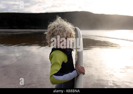 Junge Junge, bereit zum Surfen mit seinem Surfbrett am Strand das Tragen von Neoprenanzug an einem Sommertag. Stockfoto