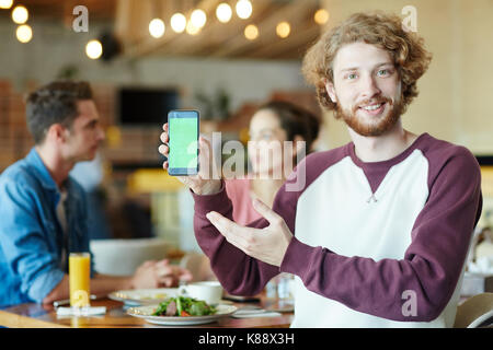 Gut aussehender bärtiger Mann mit Kamera suchen mit toothy Lächeln, während auf Leerer Bildschirm des Smartphones zeigen, seine besten Freunde hinter ihm sitzt und spricht Stockfoto