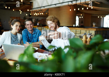 Fröhlicher junger Mitarbeiter in Freizeitmode unter selfie auf dem Smartphone beim Arbeitstreffen in der gemütlichen kleinen Kaffeehaus Stockfoto