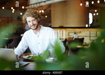 Junge Manager mit buschigen Bart an Kamera suchen und genießen Sie leckeres Essen und Arbeiten auf vielversprechende Projekt in der gemütlichen kleinen Cafe Stockfoto