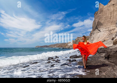 Junge Frau im roten Kleid und geht barfuß auf dem schwarzen Sand Meer Stockfoto