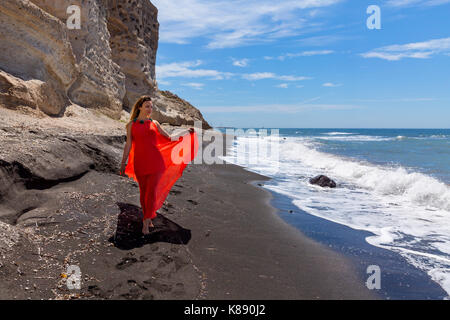 Junge Frau im roten Kleid und geht barfuß auf dem schwarzen Sand Meer Stockfoto