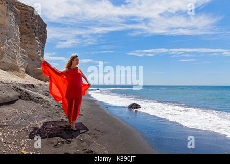 Junge Frau im roten Kleid und geht barfuß auf dem schwarzen Sand Meer Stockfoto