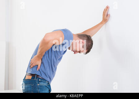 Junger Mann mit Rückenschmerzen während lehnte sich auf weiße Wand Stockfoto