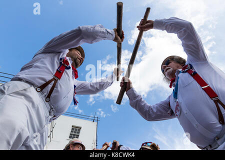 Hartley Morrismen Tänzer, in Weiß mit roten Klammern und Strohhüte gekleidet, stehend über Viewer, bashing Holzpfähle. Blauer Himmel, weiße Wolken. Stockfoto