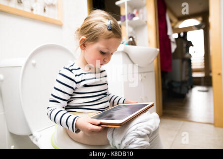 Kleines Mädchen mit Tablet auf der Toilette sitzen. Stockfoto