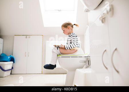 Kleines Mädchen mit Tablet auf der Toilette sitzen. Stockfoto