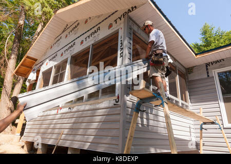 Ein Bauarbeiter, ein Stück Vinyl Siding zu einem anderen Arbeiter auf ein temporäres Gerüst in Ontario, Kanada. Stockfoto