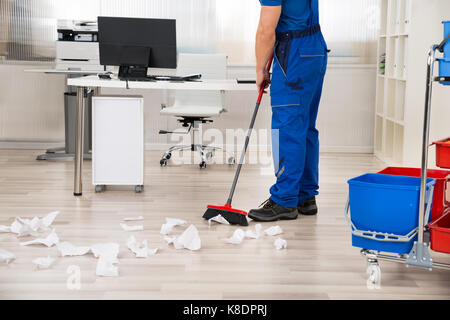 Niedrige Abschnitt der männlichen Hausmeister fegt Papiere auf dem Boden mit Besen in Office gefallen Stockfoto