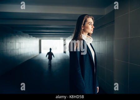 Wunderschöne einsame Frau in einem U-Bahn Tunnel mit erschrecken Silhouette auf Hintergrund. Surrealismus Konzept Stockfoto