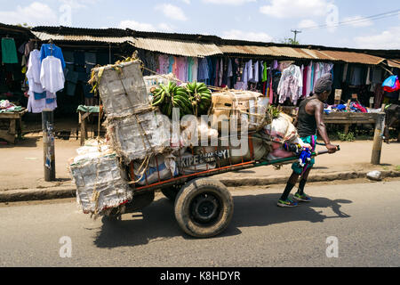 Ein Mann zieht einen Handkarren mit Gütern auf der Straße mit Kleidung Stände auf Pflaster im Hintergrund, Nairobi, Kenia Stockfoto