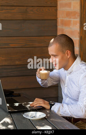 Hübscher junger Mann im Hemd sitzt, liest Nachrichten online und Kaffee zu trinken. Der Schüler sitzt in einem Café und einen Laptop verwenden Stockfoto