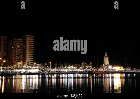 Nachtpanorama des Hafens von Malaga mit 'Farola' Leuchtturm und Meer, das die Lichter der Stadt reflektiert Stockfoto