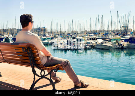 Mann sitzt und Sonnenbaden auf einer Bank der Betrachtung der Boote im Hafen von Alicante. Spanien Stockfoto