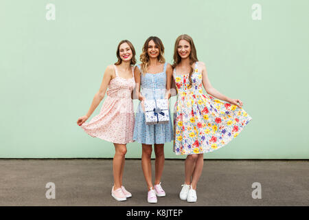 Charmante drei schöne junge erwachsene Freunde mit Kleidern und vorhanden, lächelnd und mit Blick auf die Kamera. Auf grüne Wand isoliert. Studio shot Stockfoto