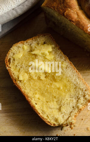 Overhead shot aus einer Scheibe frisch gebackenen, warmen Brot mit Butter schmelzen in Oberfläche. Auf Holzbrett mit Ofen Handschuhe, Laib und Krümel. Stockfoto