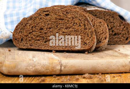 Ein Laib Schwarzbrot liegen auf einem Schneidebrett, in Scheiben schneiden. Kochen zu Hause, in der Nahaufnahme. Im Hintergrund ist eine Papierserviette. Geringe Tiefe von fi Stockfoto