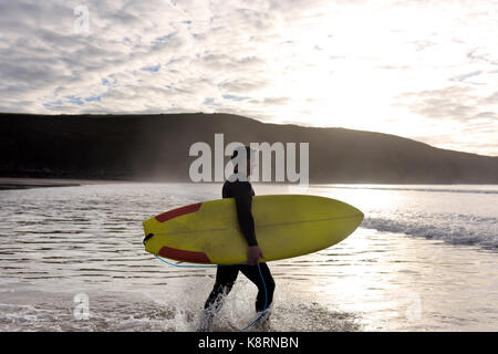 Ein Mann geht zum Meer, als er seinen nächsten Wellen am Strand betrachtet trägt einen Anzug seine eigenen Gelben Surfboard trägt. Stockfoto