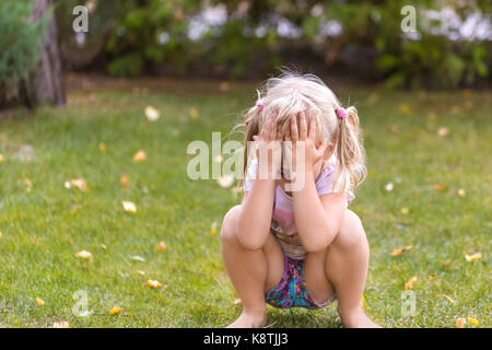 Portrait von cute adorable kleine Blonde kaukasischen Kauerndes Mädchen auf grünem Rasen im Freien, Spaß und Spielen verstecken und Spiel suchen, Ausblenden der f Stockfoto