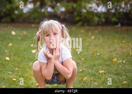 Portrait von cute adorable kleine Blonde kaukasischen Kauerndes Mädchen auf grünem Rasen im Freien, Spaß haben und fröhlich mit (nicht schlecht) f Grimasse Stockfoto