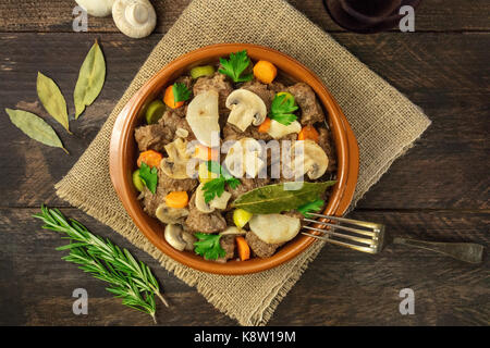 Ein Foto von einem fleischragout in einem irdenen Schüssel, der mit Zweigen von frischem Rosmarin, Lorbeer und Pilze, Schuß von oben auf einem urigen Hintergrund te Stockfoto