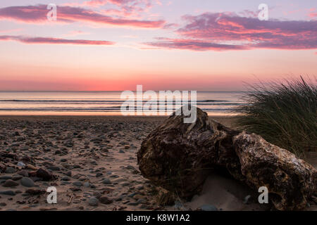 Sonnenuntergang an der Westküste des Vereinigten Königreichs, North Wales. Rock im Vordergrund, Sand und Kies, Gras nach rechts des Rahmens. Ruhiges Meer. Überwiegend klar Stockfoto