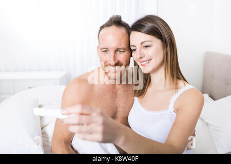 Fröhliche Paare herausfinden, Ergebnisse einer Schwangerschaft Test sitzen auf dem Bett Stockfoto