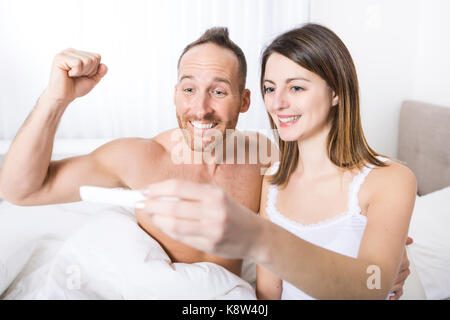 Fröhliche Paare herausfinden, Ergebnisse einer Schwangerschaft Test sitzen auf dem Bett Stockfoto
