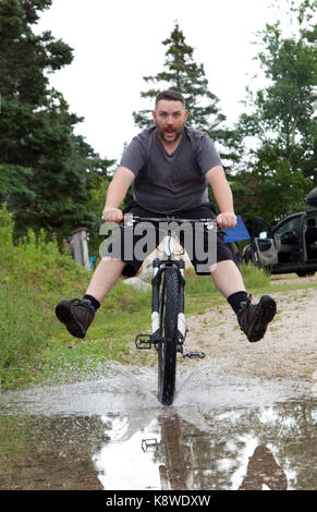 Ein Mann mit aufgeregten Ausdruck fährt mit dem Fahrrad durch eine große Pfütze Stockfoto