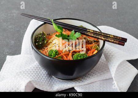 Reisnudeln mit Karotten, Brokkoli und Champignons in einer schwarzen Schale auf einem schwarzen Hintergrund abstrakt. Asiatische Küche. Gesunde Ernährung Stockfoto