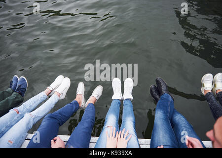 Jugendliche Freunde sitzen auf der Brücke über den Fluss, Lifestyle, Füße über blaue Wasser Stockfoto