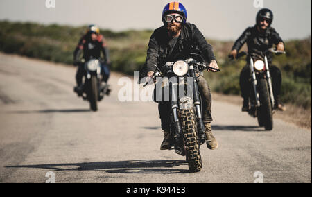 Drei Männer mit offenem Gesicht Schutzhelme und -brillen, Cafe Racer Motorrad entlang der Landstraße. Stockfoto