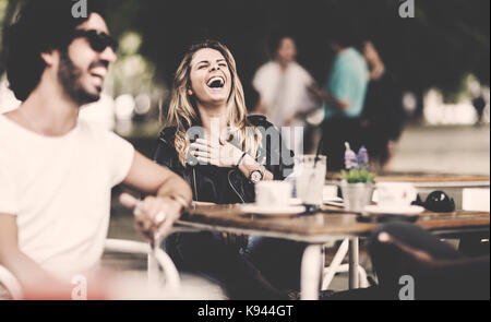 Bärtiger Mann mit Sonnenbrille und Frau mit langen blonden Haaren im Freien sitzen an einem Tisch in einem Cafe, Lachen. Stockfoto