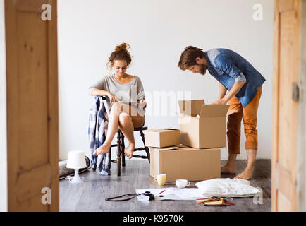 Junges Ehepaar Umzug in ein neues Haus, Frau mit Tablet sitzt auf einem Stuhl, Mann auspacken Dinge aus einem Karton. Stockfoto