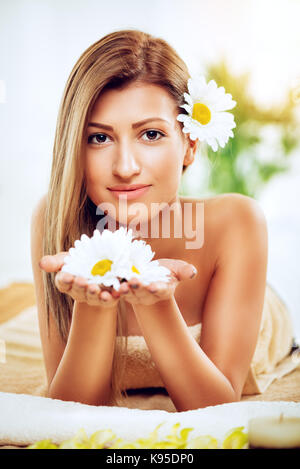 Nette junge lächelnde Frau genießen Sie während einer skincare Behandlung in einem Spa. Sie schaut an Kamera und halten weiße Blumen in den Händen. Stockfoto