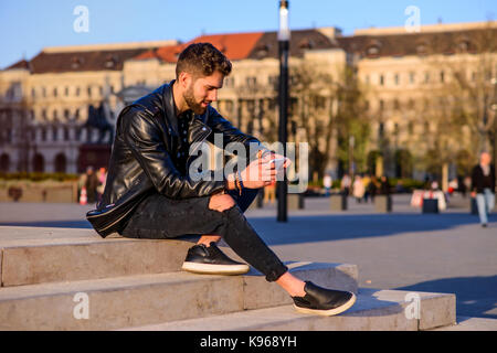 Ein hübscher junger hipster Mann sitzt auf der Treppe auf einem Platz mit seinem Smartphone Stockfoto