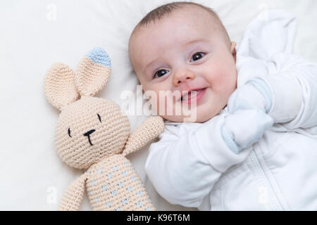 BABY MIT SCHNULLER. Kleiner Junge, der glücklich und lächelnd auf dem Bett ruht, mit einem Hasen-Dummy aus Häkelgewebe zur Seite. Draufsicht. Stockfoto