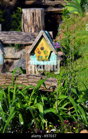 Rustikaler Zaun ist mit einem bunt bemalten Vogelhaus ziert. Blumen blühen im Garten. Stockfoto