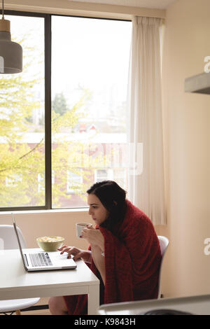 Frau eingewickelt in Decke Kaffee, während Sie laptop von Fenster zu Hause Stockfoto