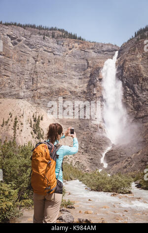 Weibliche Wanderer nehmen Bild von Wasserfall mit Handy an einem sonnigen Tag Stockfoto