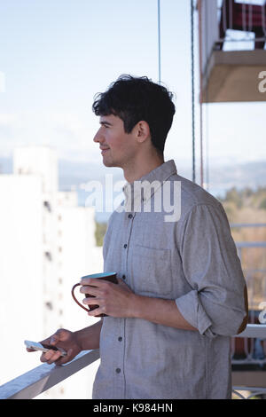 Lächelnder Mann mit Handy und in Tasse Kaffee auf dem Balkon Stockfoto